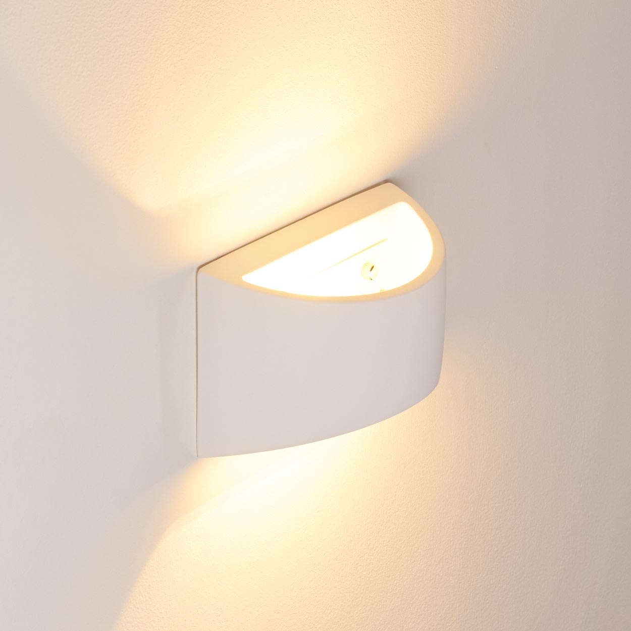 Eine weiße, gebogene Wandleuchte ist an einer weißen Wand montiert. Die Lampe strahlt ein warmes, gelbliches Licht nach oben und unten aus.