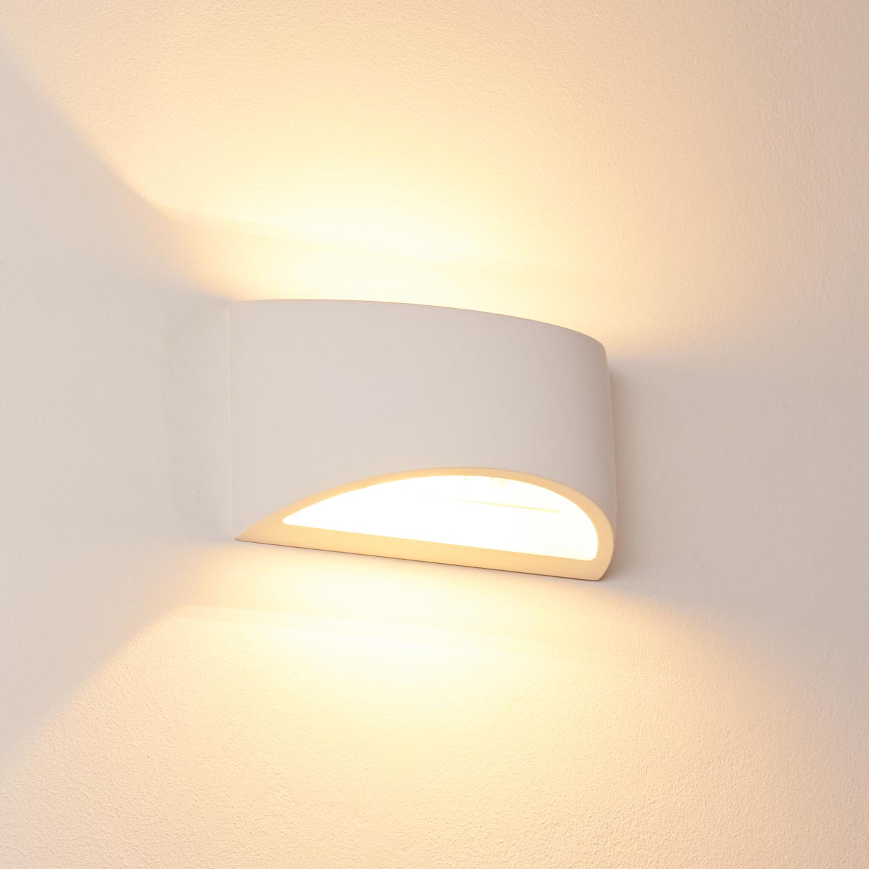 Eine weiße Wandleuchte mit geschwungenem Design. Sie strahlt ein warmes, helles Licht aus und wirft Schatten an die Wand.