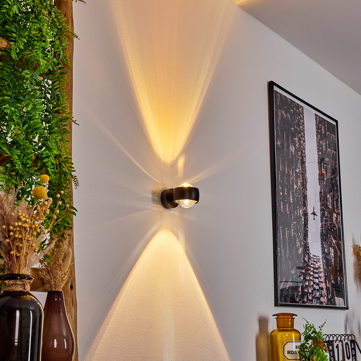 Moderne schwarze Wandleuchte mit Lichtstrahlen. Ein gerahmtes Bild, Pflanzen und Vasen sind an einer weißen Wand zu sehen.
