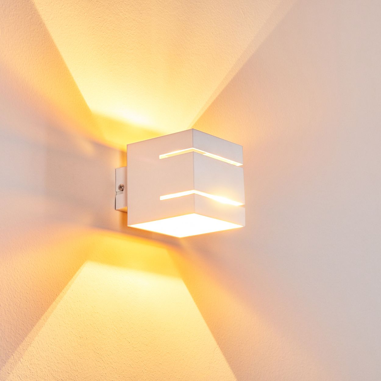 Eine weiße, würfelförmige Wandleuchte ist an einer Wand montiert. Die Lampe strahlt warmes Licht aus und wirft einen Schein. Die Wand ist hell und neutral.