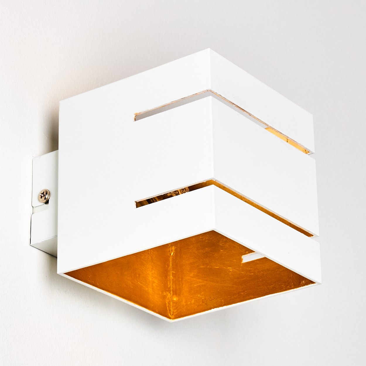 Eine weiße, würfelförmige Wandleuchte mit goldfarbenem Innenraum. Sie ist an einer weißen Wand montiert. Die Lampe hat ausgeschnittene Linien.