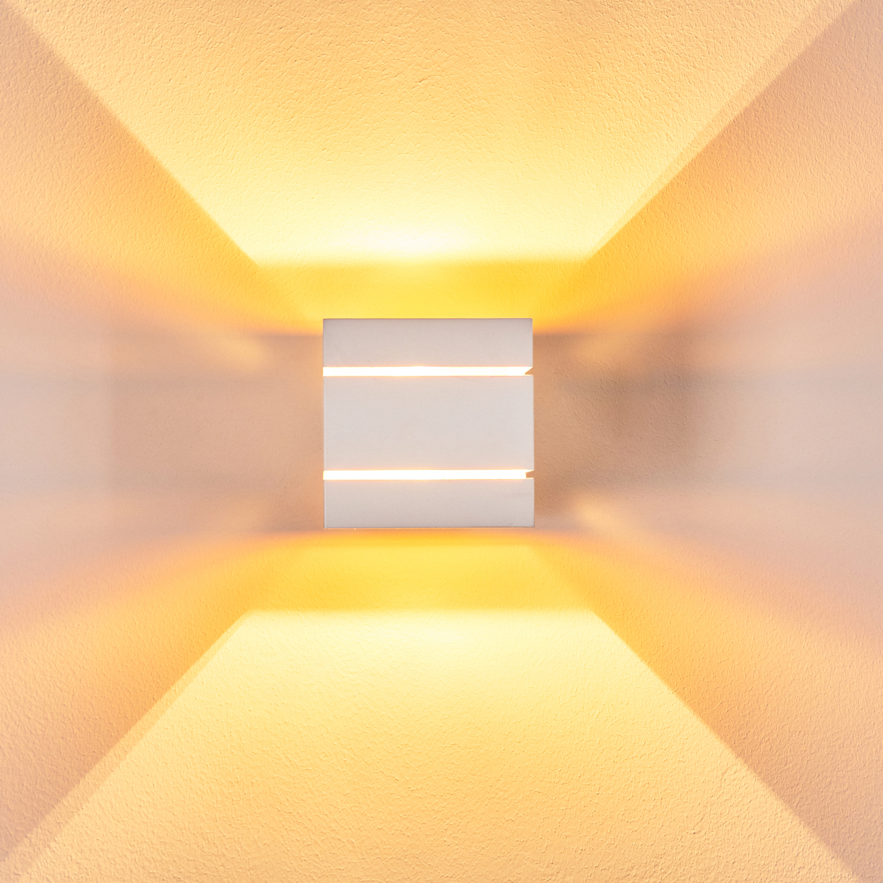 Eine weiße Wandleuchte mit drei horizontalen Schlitzen, die Licht emittieren. Das Licht wirft einen gelben Schein auf die Wand.