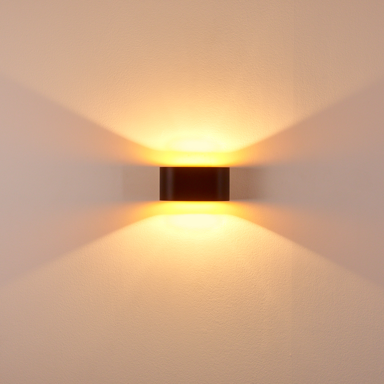 Eine dunkle, rechteckige Wandleuchte ist an einer strukturierten, hellfarbigen Wand angebracht. Die Lampe strahlt warmes Licht nach oben und unten aus.