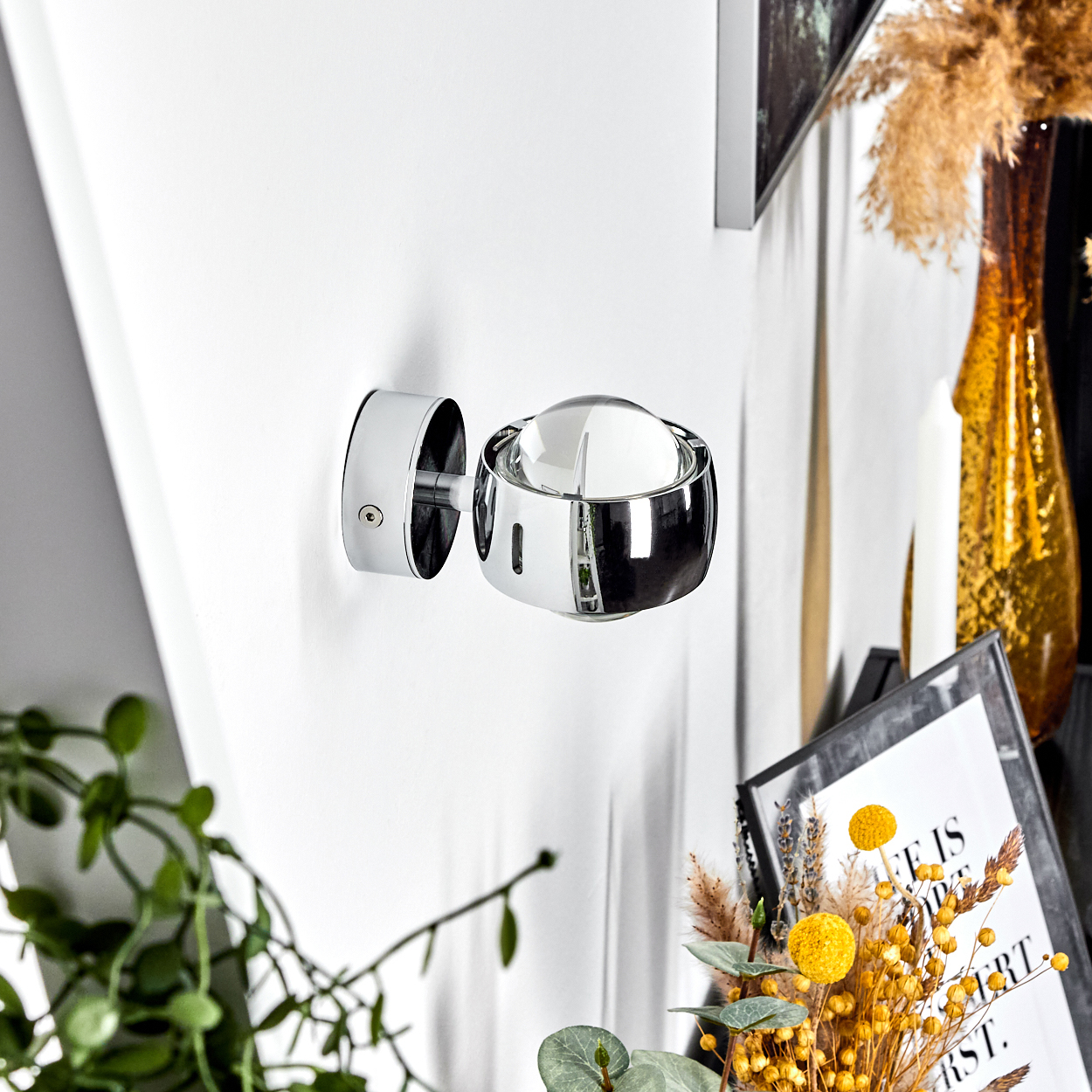 Eine verchromte Wandlampe mit Glaskuppel ist an einer weißen Wand befestigt. Eine Vase, Blumen und gerahmte Kunst sind sichtbar.
