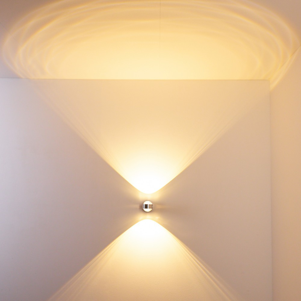 Eine Wandleuchte mit zwei Lichtstrahlen. Das Licht wirft ein V auf die Wand. Die Wand ist weiß.
