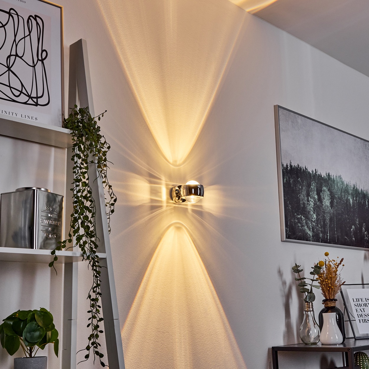 Eine moderne Wandlampe wirft Lichtmuster auf eine weiße Wand. Dekorationen umfassen Pflanzen, Kunst und einen kleinen Tisch mit Vasen.