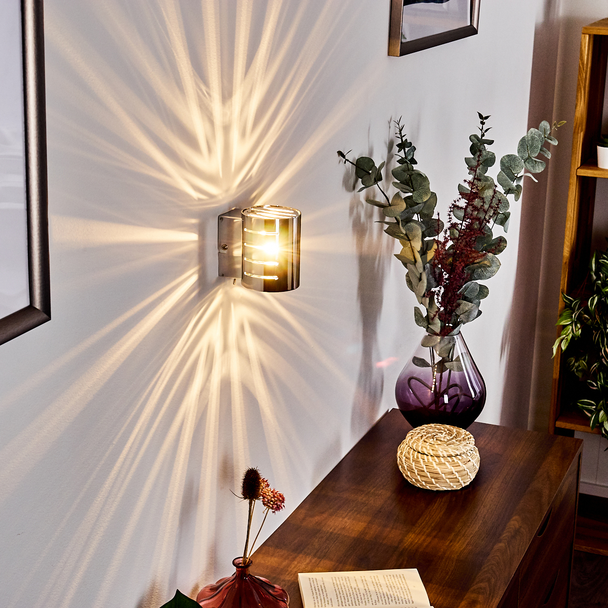 Eine Wandlampe wirft Lichtmuster an die Wand. Eine Vase mit Blumen und ein Weidenkorb stehen auf einem Holztisch. Ein Buch ist offen.