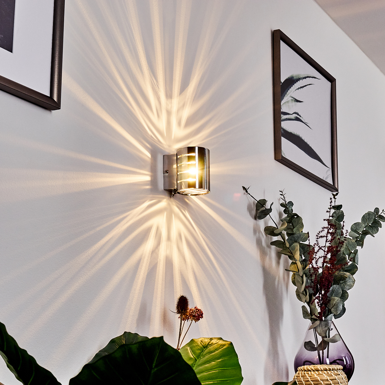Eine Wandleuchte wirft Lichtmuster. Ein Bilderrahmen hängt an der Wand, und eine Vase mit Pflanzen befindet sich im Vordergrund.