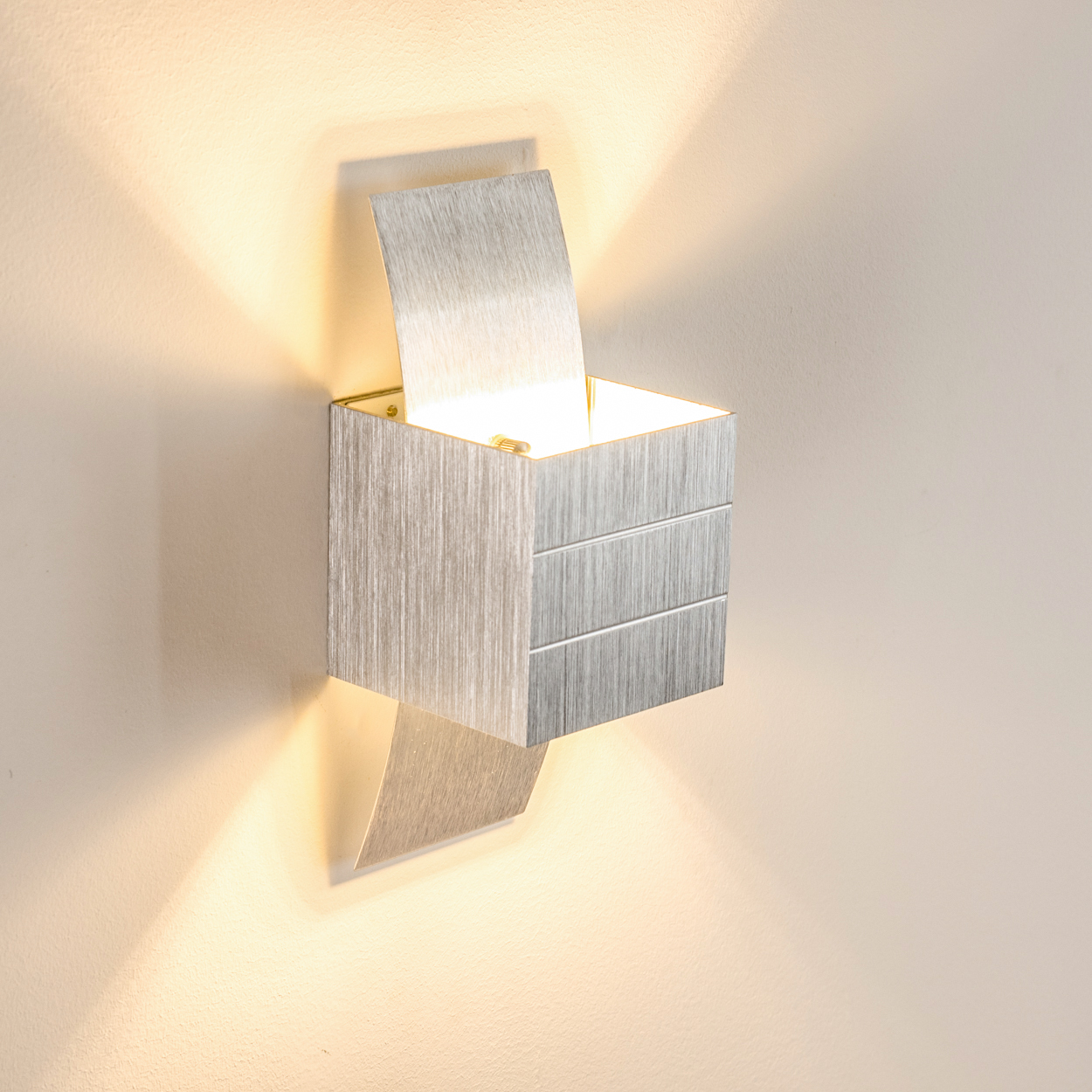 Eine moderne silberne Wandlampe mit offenem Deckel, die ein warmes Licht auf eine helle Wand wirft. Die Lampe hat ein einzigartiges Design.