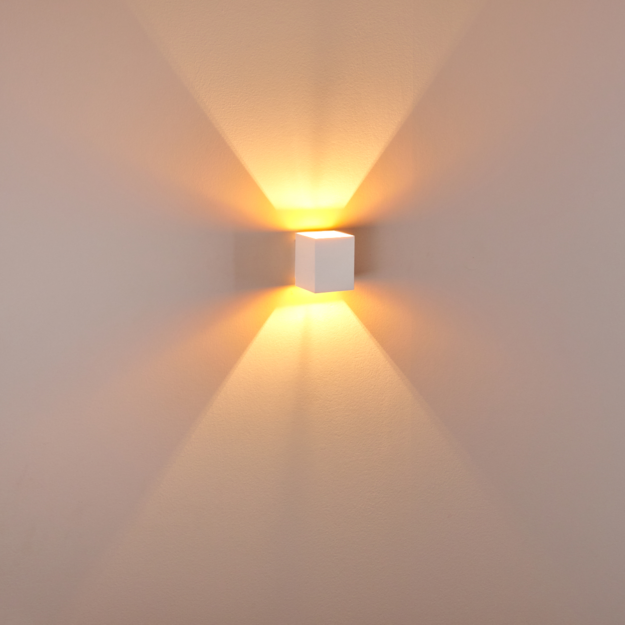 Ein weißer, würfelförmiger Wandstrahler strahlt hellgelbes Licht aus. Das Licht wirft einen fächerförmigen Schatten auf die hellbraune Wand.