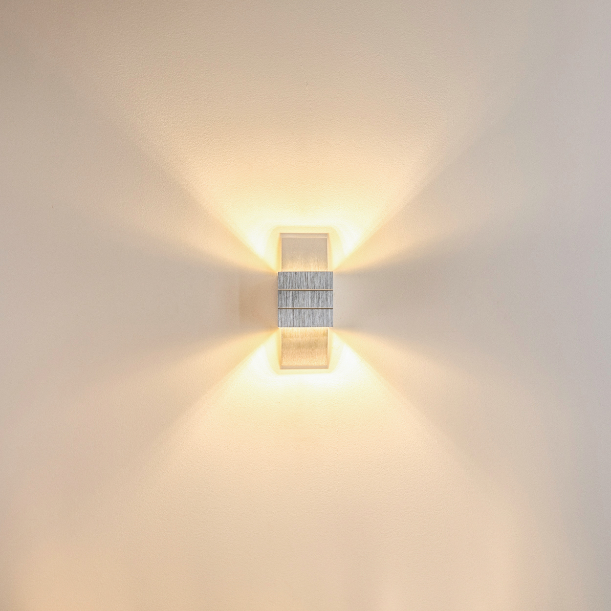 Eine moderne Wandlampe mit metallischer Mitte und Glasplatten, die warmes Licht ausstrahlt. Sie ist an einer beigefarbenen Wand befestigt.