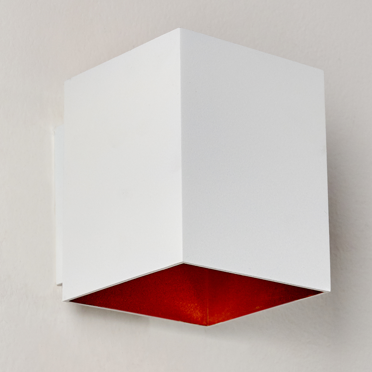 Eine weiße, würfelförmige Lampe mit rotem Innenraum ist an einer weißen Wand befestigt. Die Lampe ist ein modernes Design.