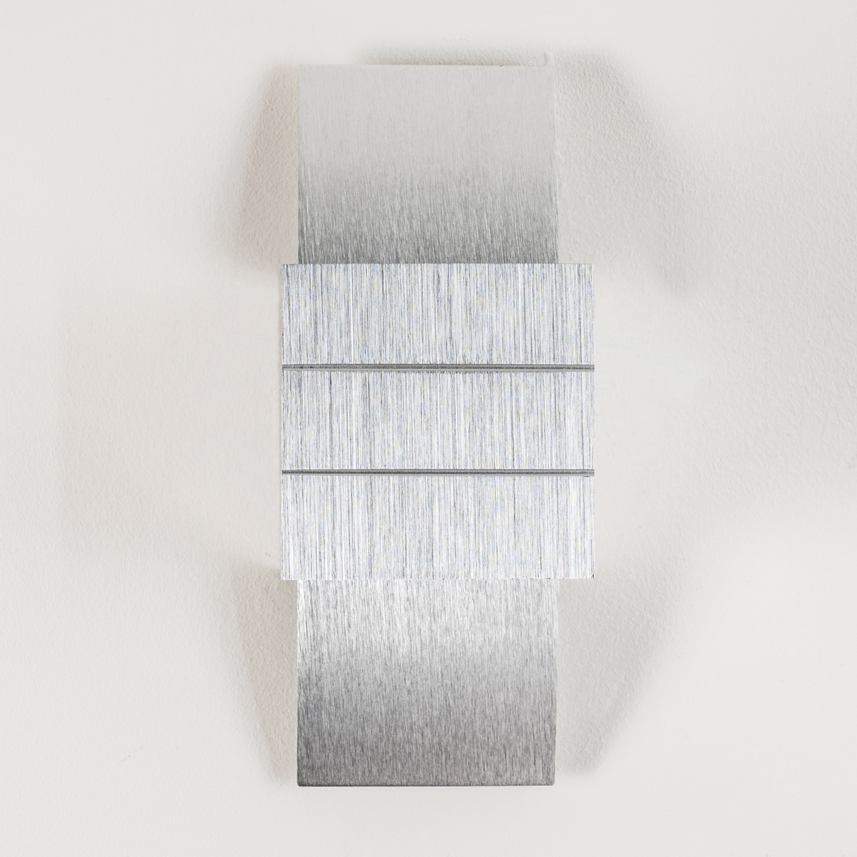 Eine metallische Skulptur aus drei rechteckigen Teilen mit gebürsteter Metallstruktur, die an einer weißen Wand befestigt ist.