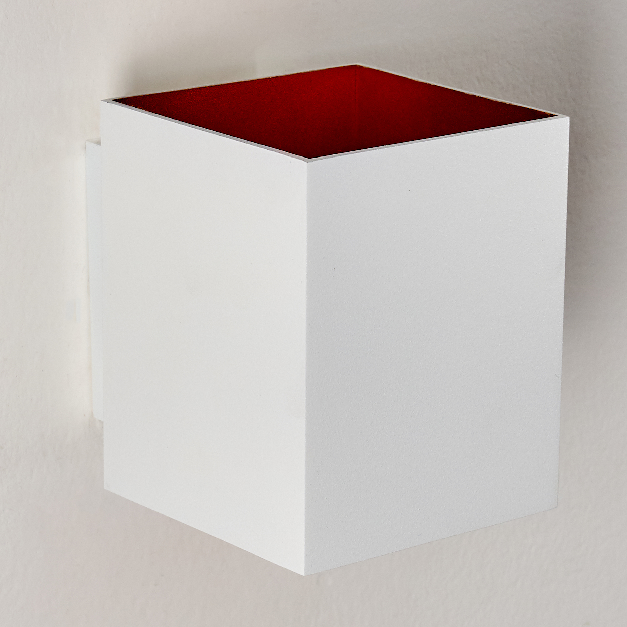 Ein weißer Würfel mit rotem Innenraum ist an einer weißen Wand befestigt. Der Würfel hat eine offene Oberseite. Die Wand ist strukturiert.