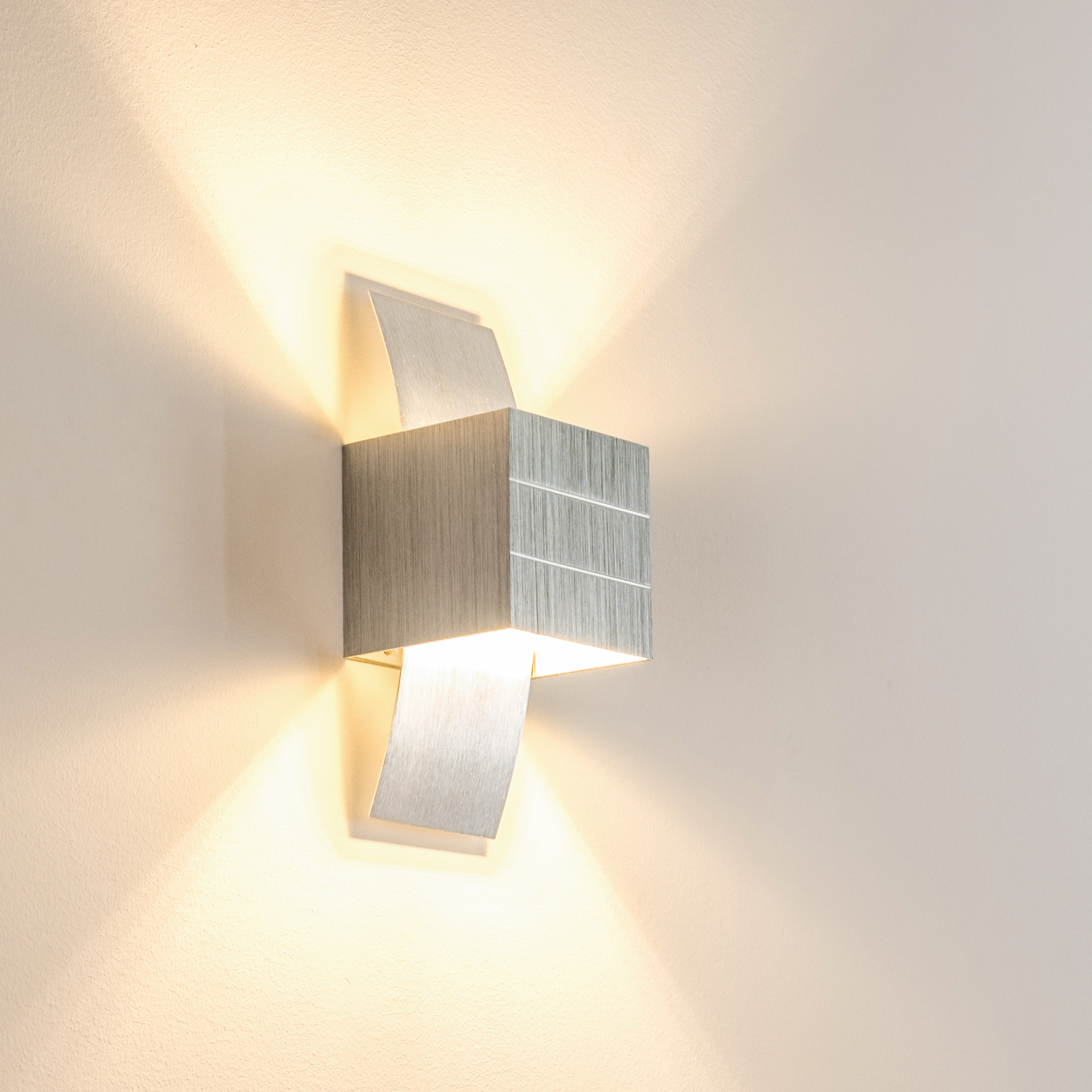 Eine moderne, metallische Wandleuchte strahlt warmes Licht auf eine beige Wand. Die Lampe hat eine Würfelform mit zwei Klappen.