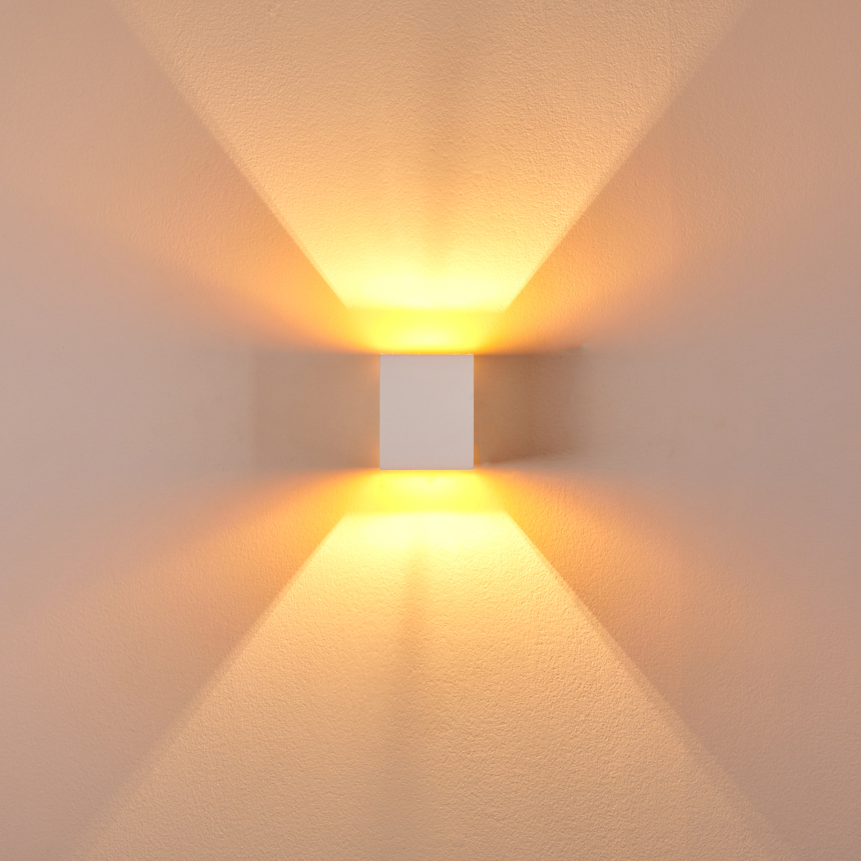 Eine quadratische, weiße Wandleuchte strahlt warmes Licht nach oben und unten aus. Das Licht erzeugt dreieckige Schatten an der Wand.
