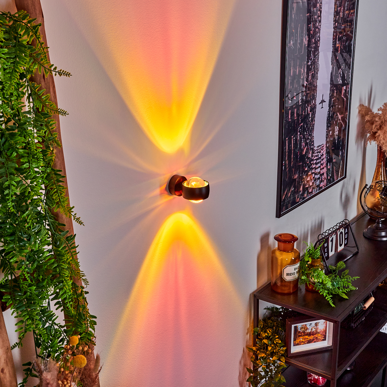 Eine Wandleuchte projiziert orange und gelbe Strahlen. Auf einem Regal befinden sich ein Bild, Deko und Pflanzen. Links ist eine Holzleiter mit Grün.