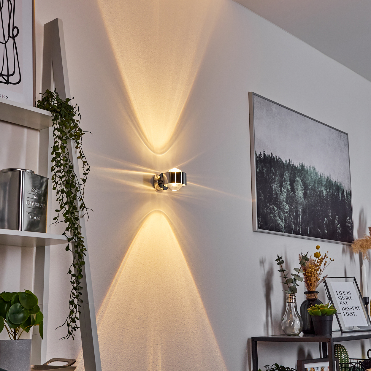 Eine Wandleuchte wirft Licht nach oben und unten. An der Wand befinden sich ein Bild und ein Regal mit Dekoration. Eine Pflanze hängt von einer Leiter.