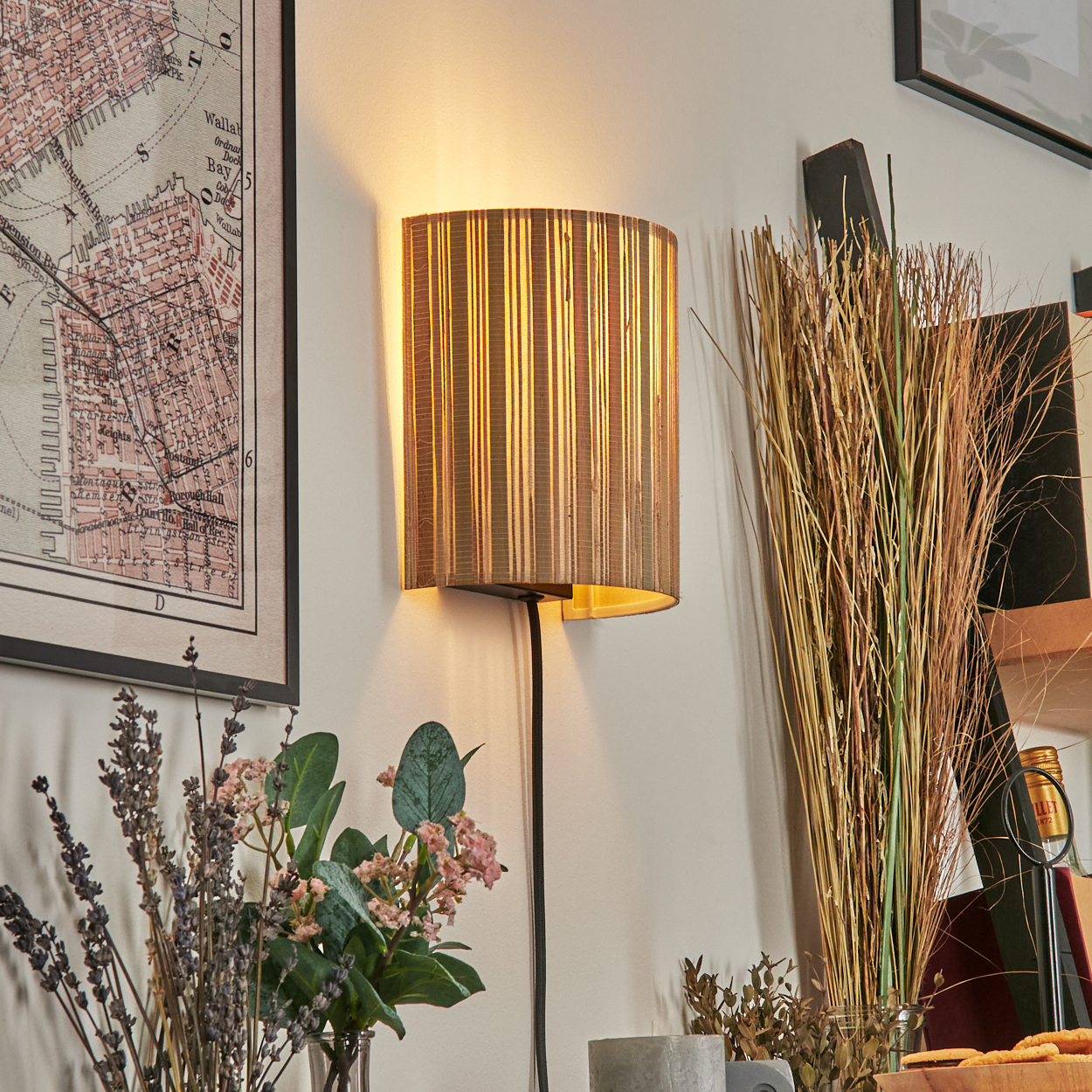 Eine Wandlampe mit gestreiftem Schirm leuchtet. Eine Karte, Blumen und Ziergräser sind in der Nähe. Das Kabel der Lampe hängt herunter.