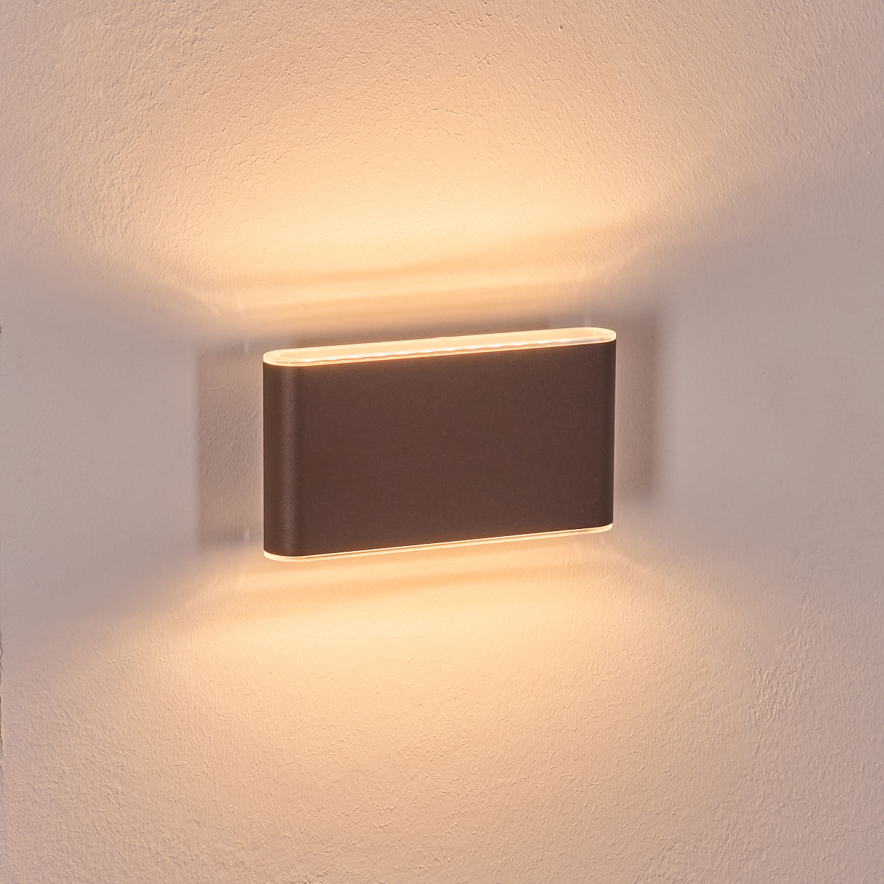 Eine dunkelbraune, rechteckige Wandleuchte ist an einer weißen, strukturierten Wand angebracht. Sie strahlt ein weiches, warmes Licht aus.
