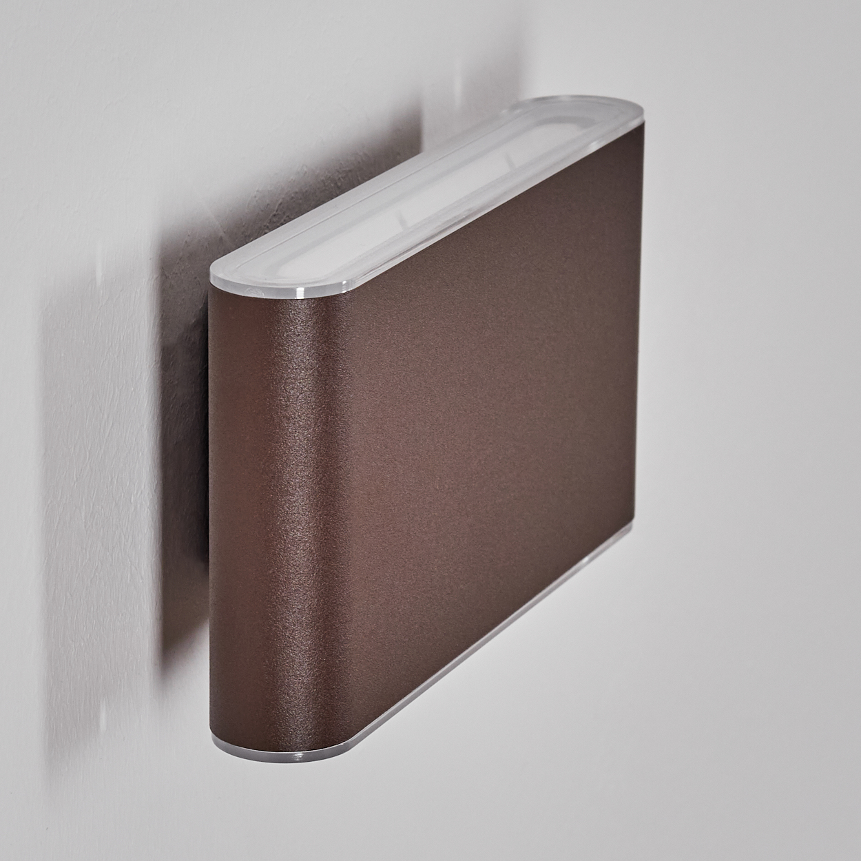 Eine braune, gebogene Wandleuchte mit weißem Oberteil und silberner Blende ist an einer weißen Wand montiert. Der Schatten der Lampe ist sichtbar.