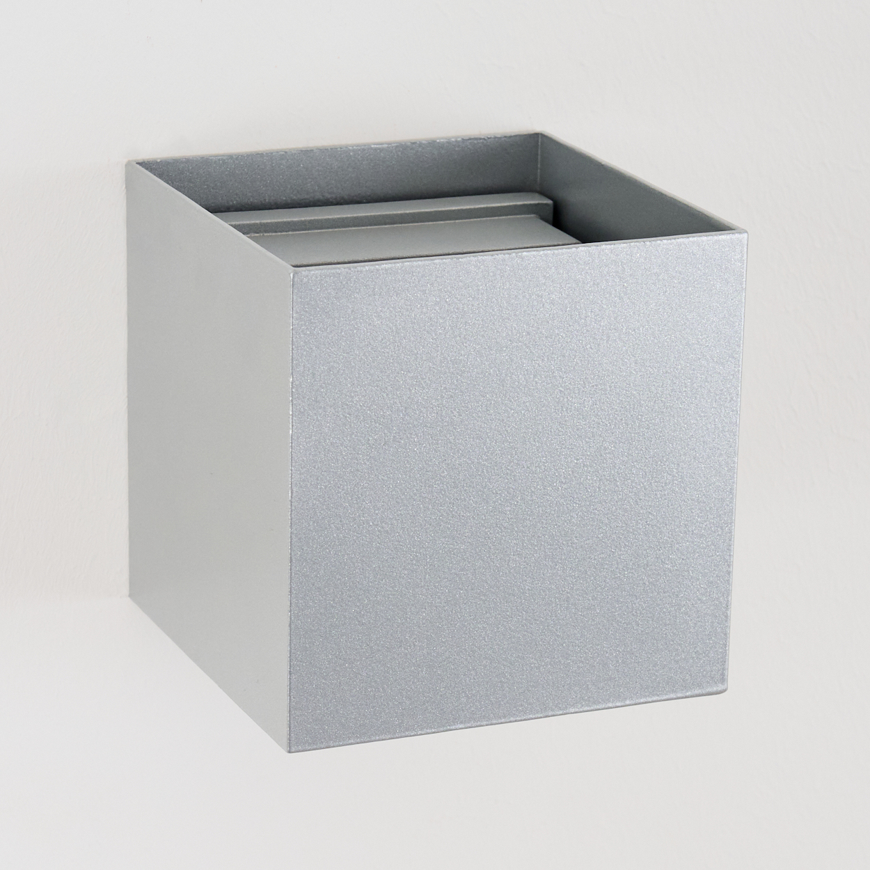 Eine quadratische, silberne Metallbox ist an einer weißen Wand befestigt. Die Box hat eine offene Oberseite mit einer kleineren Box darin.