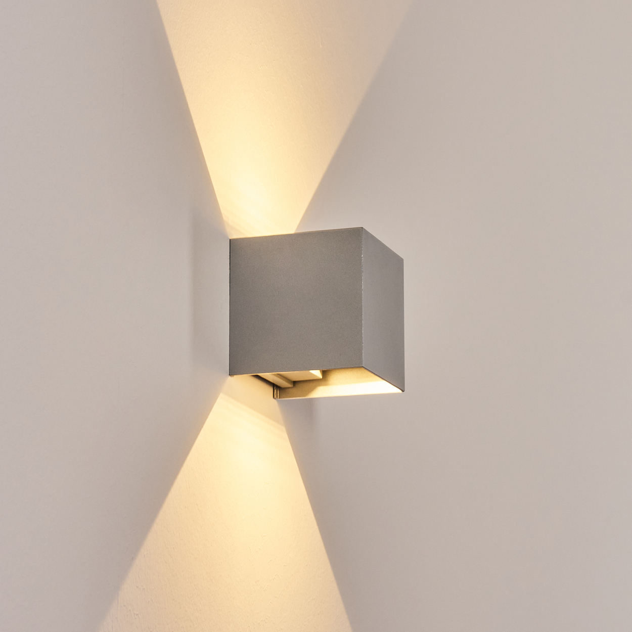 Eine graue, würfelförmige Wandleuchte ist an einer weißen Wand montiert. Die Lampe strahlt nach oben und unten und erzeugt helle Muster.