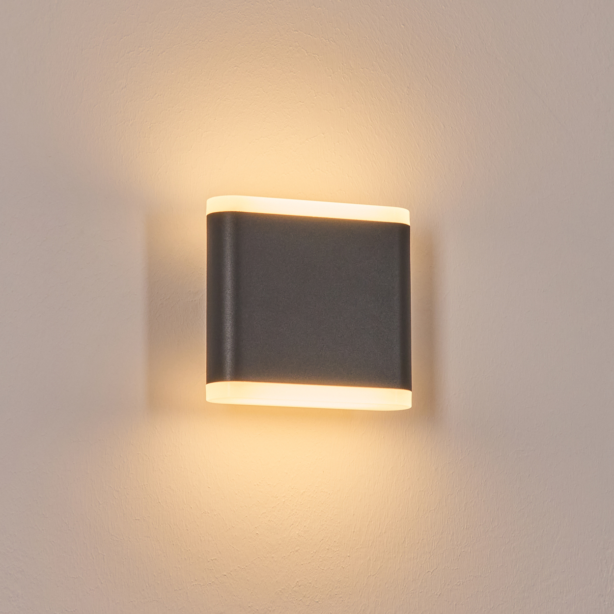 Eine moderne, rechteckige Wandleuchte mit dunkelgrauem Körper strahlt ein warmes, weiches Licht aus. Sie ist an einer strukturierten, hellbeigen Wand angebracht.
