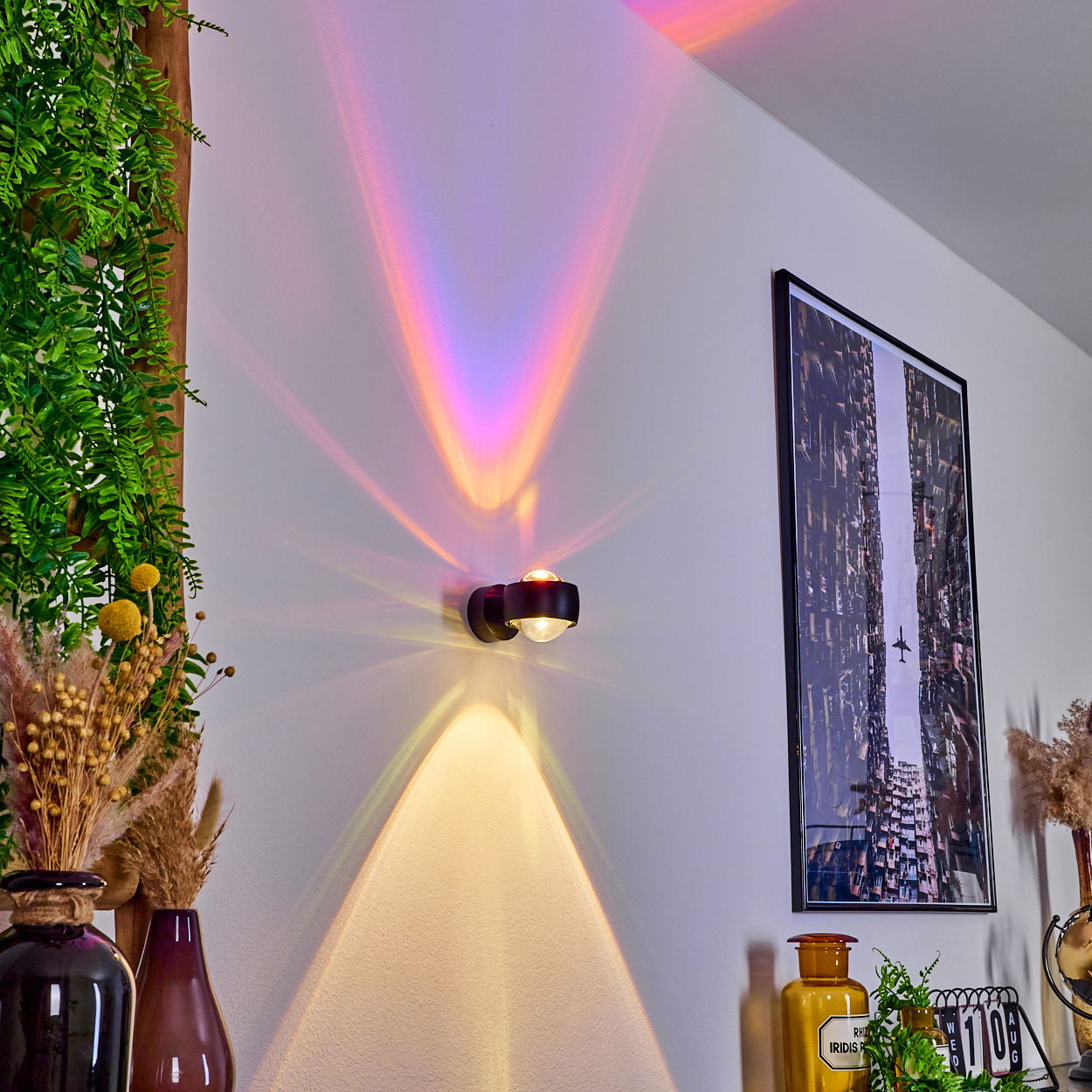 Eine Wand mit einer schwarzen Lampe, die farbenfrohe Lichtmuster projiziert. Ein Bild einer Stadt und einige Vasen mit Pflanzen sind sichtbar.