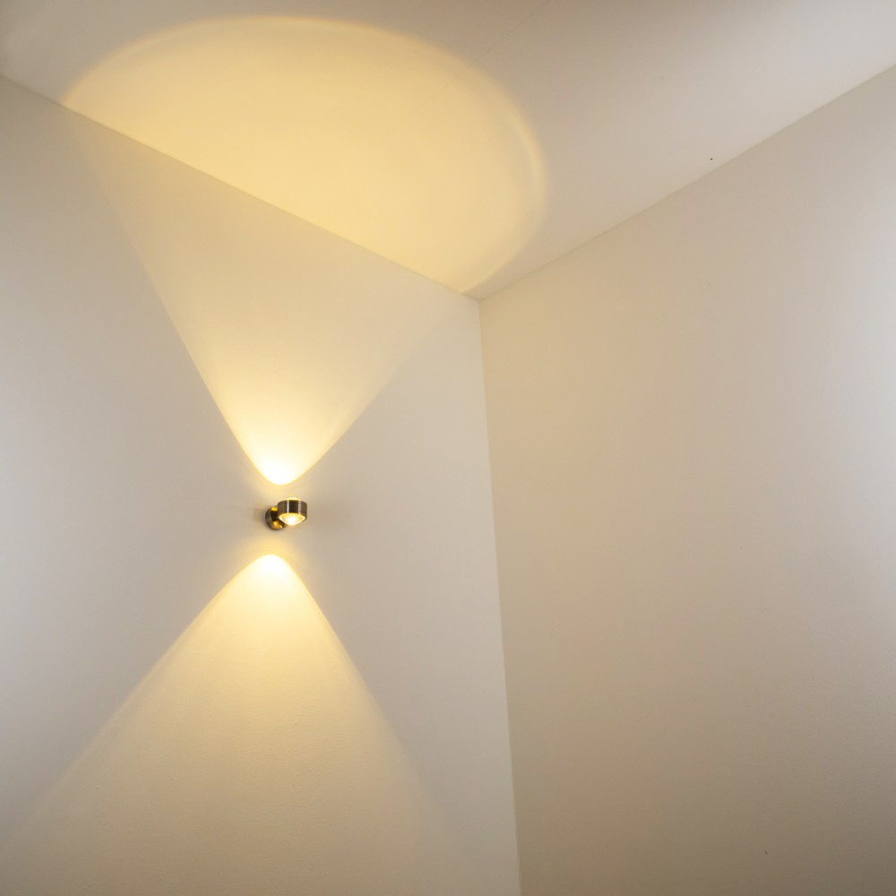 Eine Wandleuchte wirft zwei Lichtstrahlen nach oben und unten auf eine weiße Wand. Die Decke hat ein kreisförmiges Lichtmuster.