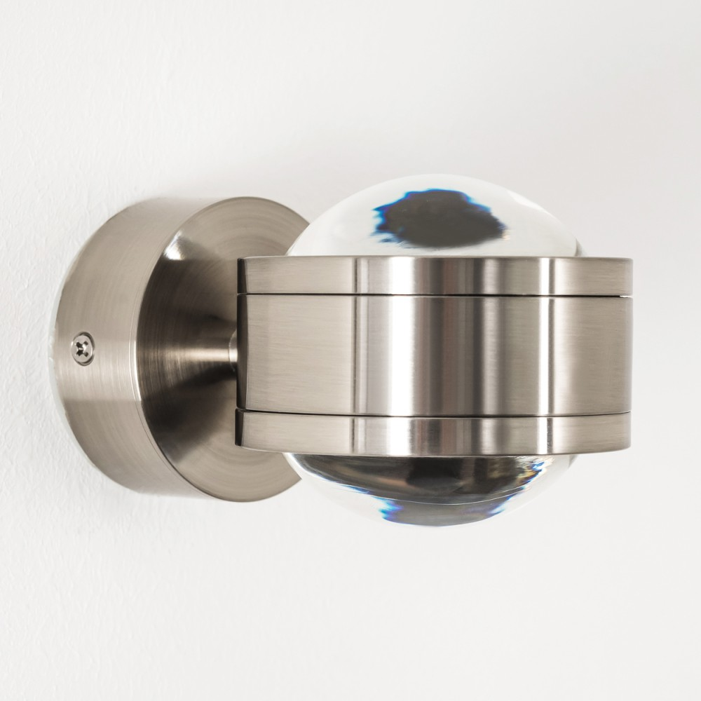 Eine moderne, metallische Wandleuchte. Sie hat einen abgerundeten Glasschirm und einen zylindrischen Metallrahmen. Die Leuchte ist an einer weißen Wand befestigt.
