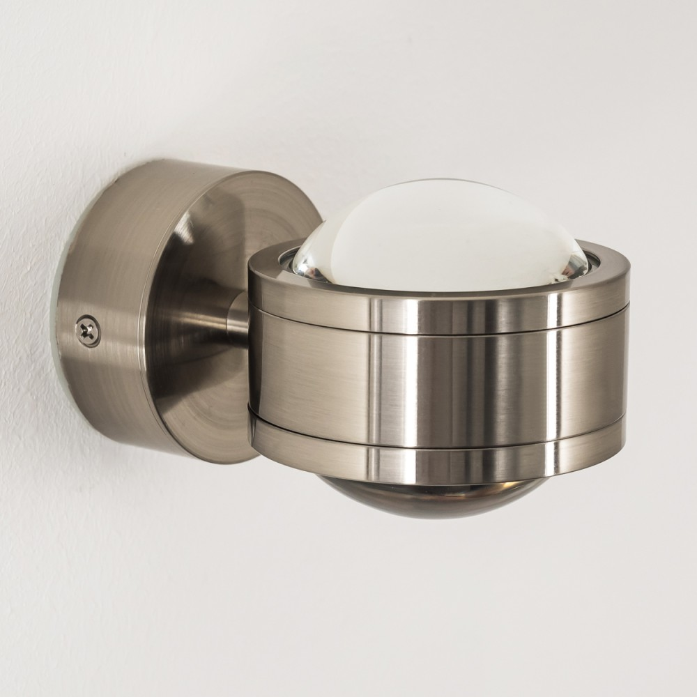 Eine metallische Wandlampe mit rundem Sockel und einer halbkugelförmigen Glasabdeckung, an einer weißen Wand befestigt.