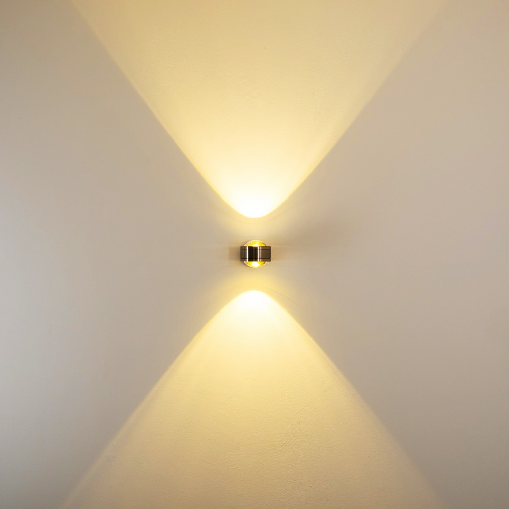 Eine Wandleuchte strahlt Licht nach oben und unten aus. Die Wand ist hellbeige. Die Leuchte ist metallisch.