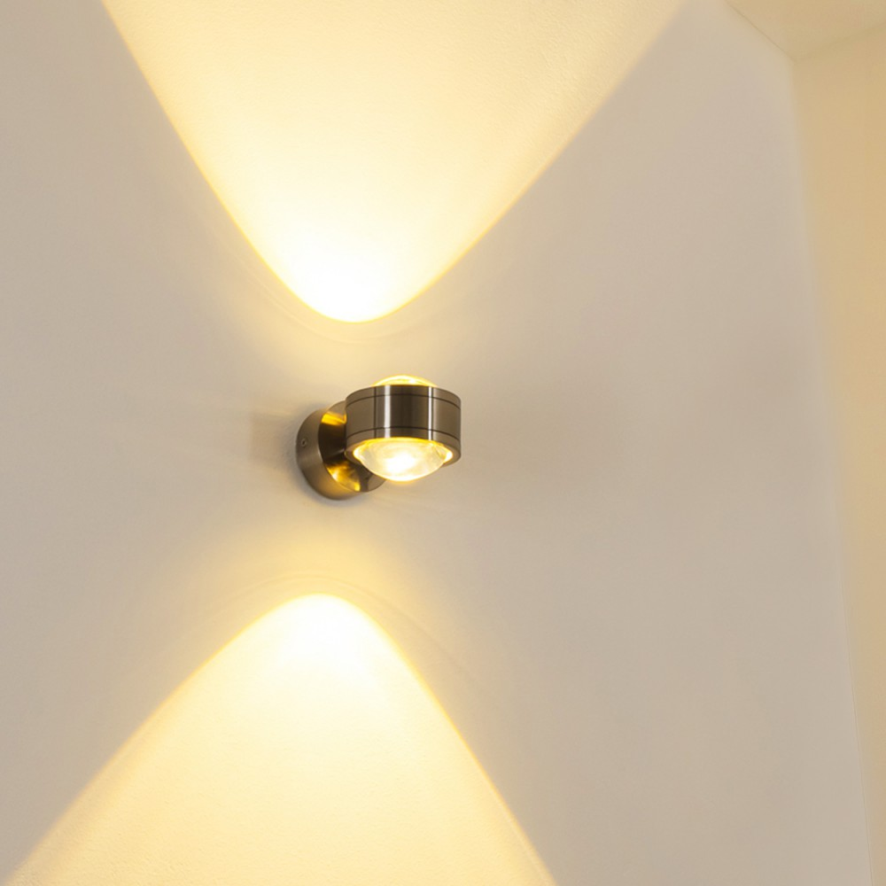 Eine moderne Wandleuchte beleuchtet eine weiße Wand. Das Licht wirft zwei sich gegenüberliegende, fächerförmige Strahlen.