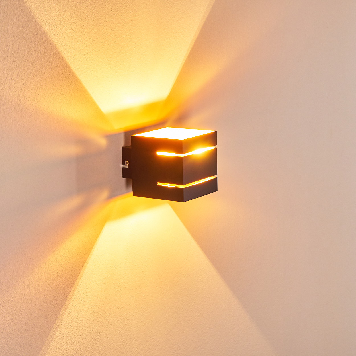 Eine moderne schwarze Wandleuchte strahlt warmes Licht aus. Das Licht erzeugt dreieckige Muster an der beigen Wand, über und unter der Lampe.