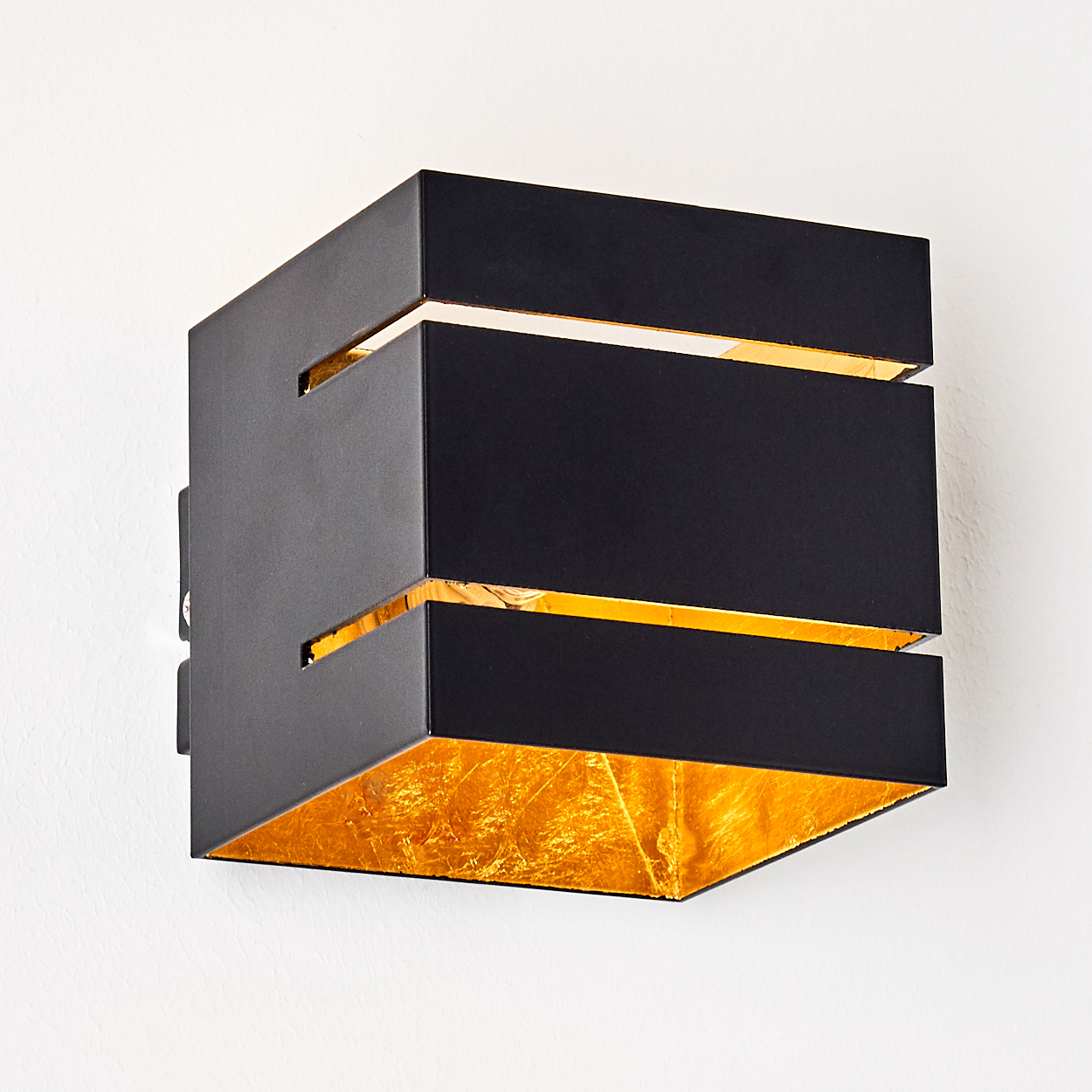 Eine moderne schwarze Wandlampe mit Blattgold im Inneren. Die Lampe hat eine Würfelform mit horizontalen Schlitzen, montiert an einer weißen Wand.