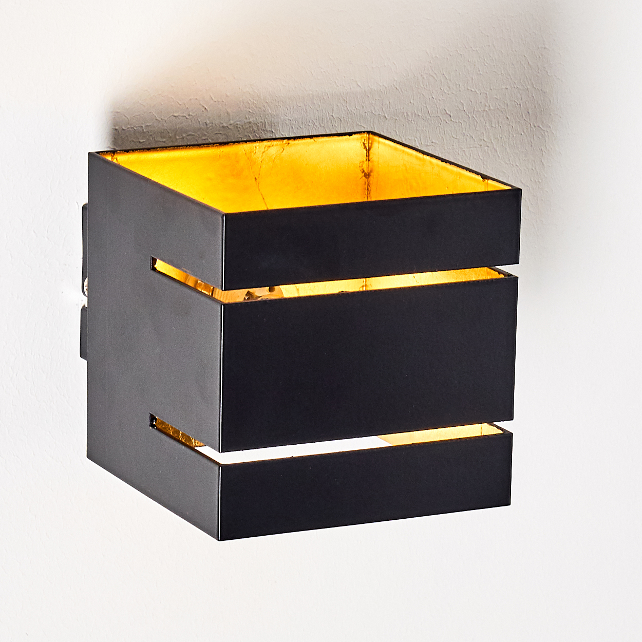 Schwarze Wandleuchte mit goldenem Innenraum und Schlitzen, an einer weißen Wand. Die Lampe hat eine kubische Form.