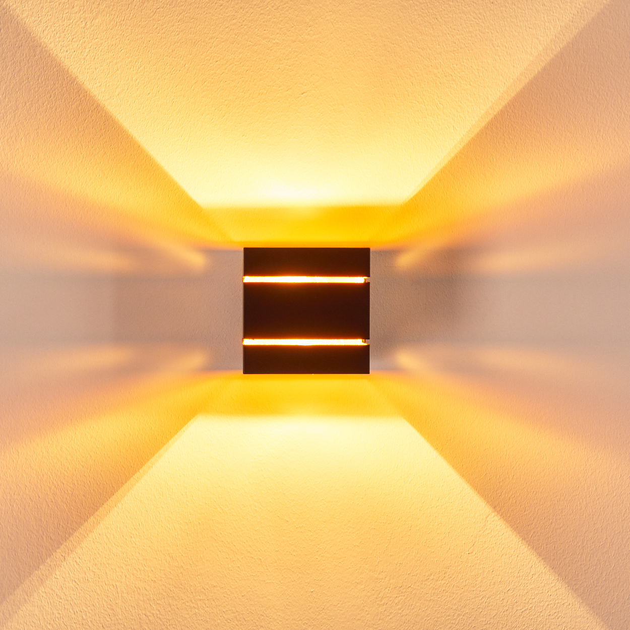 Eine dunkle, rechteckige Wandleuchte wirft starkes Licht nach oben und unten auf eine strukturierte Wand. Das Licht ist warm.