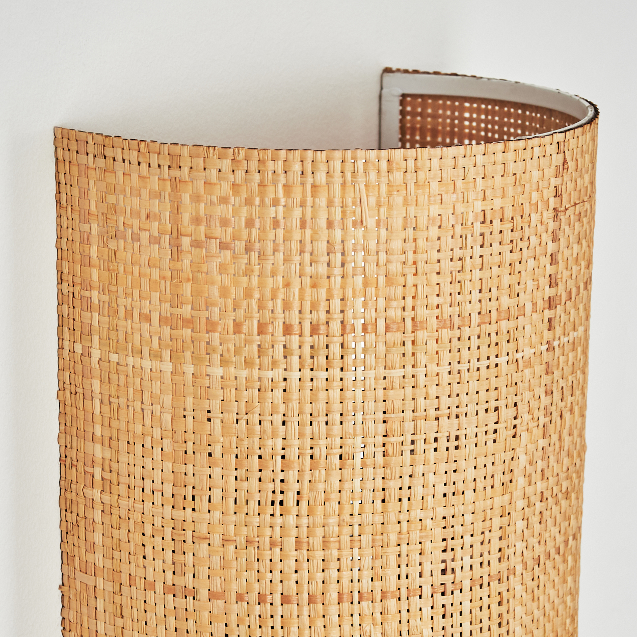 Eine gewebte, gebogene Wandlampe aus hellbraunem Rattan ist an einer weißen Wand befestigt. Die Lampe hat eine zylindrische Form.