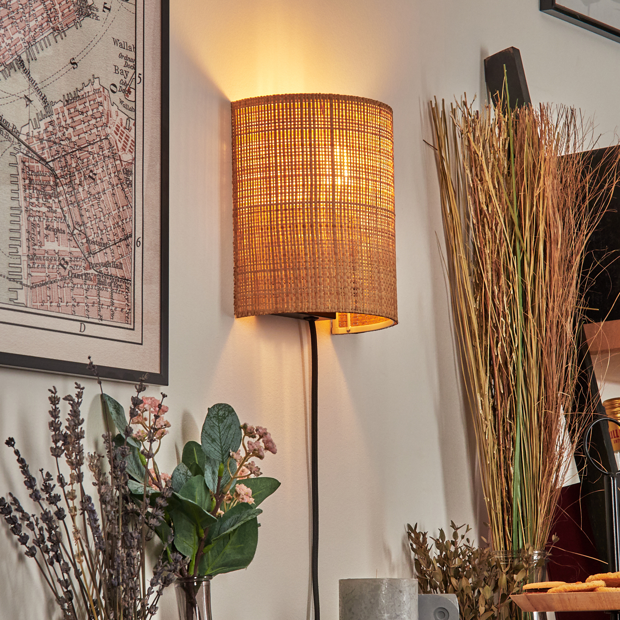 Eine gewebte Lampe beleuchtet eine Wand mit Karte, Blumen und Gräsern. Eine Kerze und Kekse stehen auf einem Tisch.