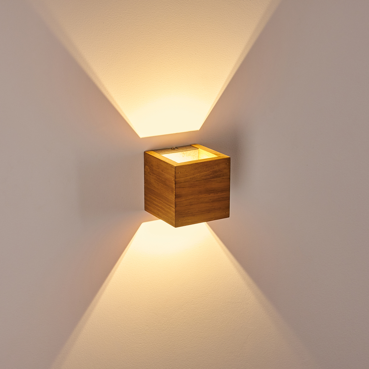 Eine quadratische Wandleuchte aus Holz strahlt Licht nach oben und unten aus. Das Licht erzeugt zwei dreieckige Muster an der Wand.
