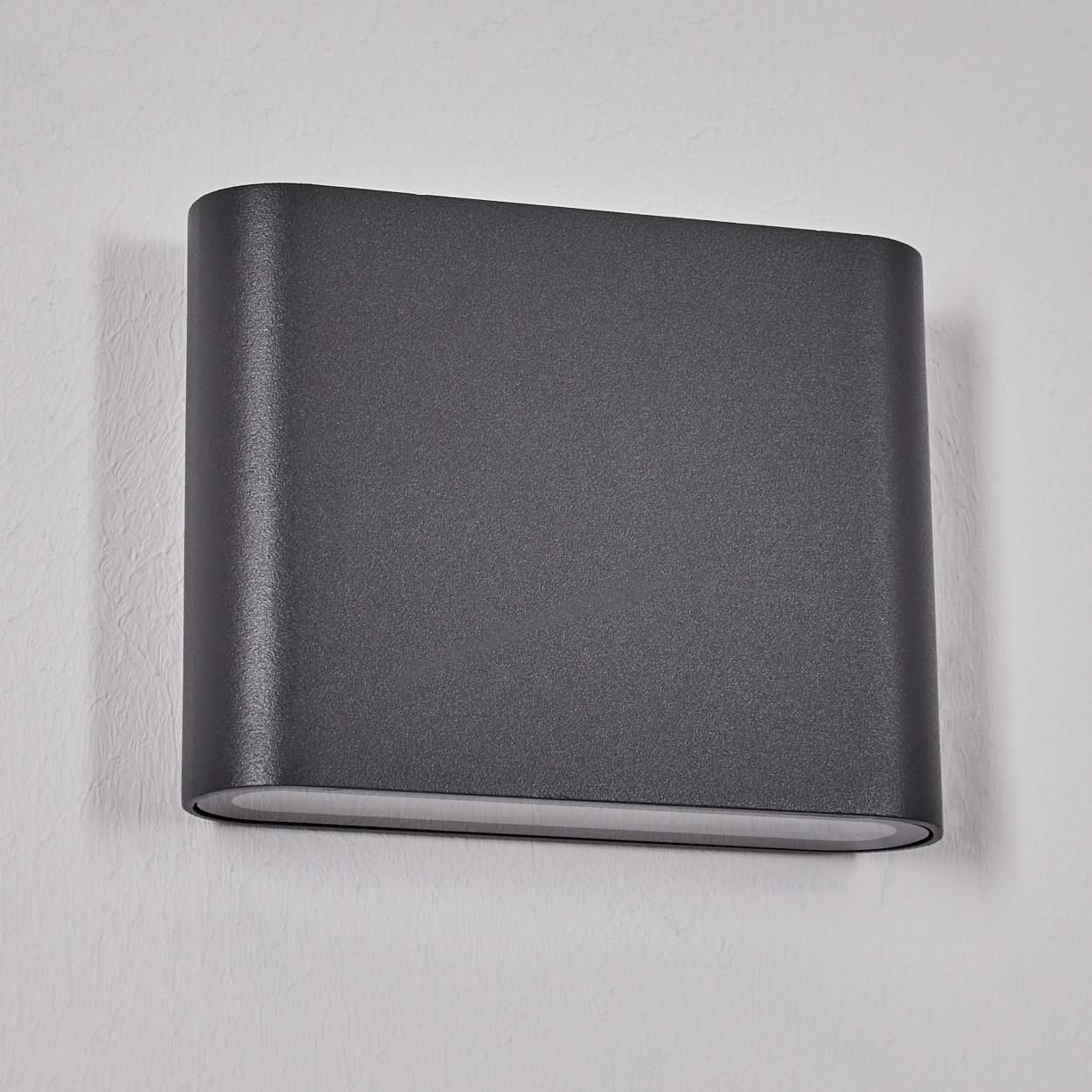 Eine dunkelgraue, rechteckige, an der Wand befestigte Leuchte mit abgerundeten Kanten. Sie befindet sich an einer strukturierten weißen Wand.