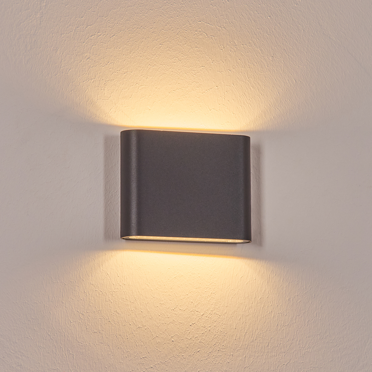 Eine dunkelgraue, rechteckige Wandleuchte ist an einer strukturierten weißen Wand befestigt. Die Lampe strahlt warmes Licht aus.