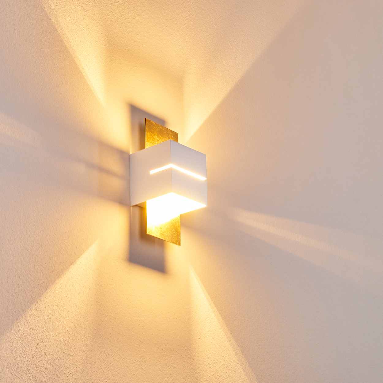 Eine moderne Wandleuchte mit weißem Quader-Schirm und goldenen Akzenten, die Licht und Schatten wirft.