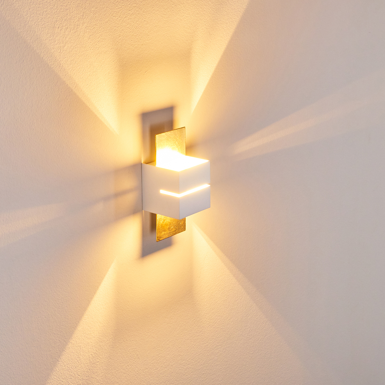 Eine moderne Wandleuchte mit weißem kubischem Schirm und goldenen Akzenten. Das Licht wirft einen hellen Schein an die Wand.