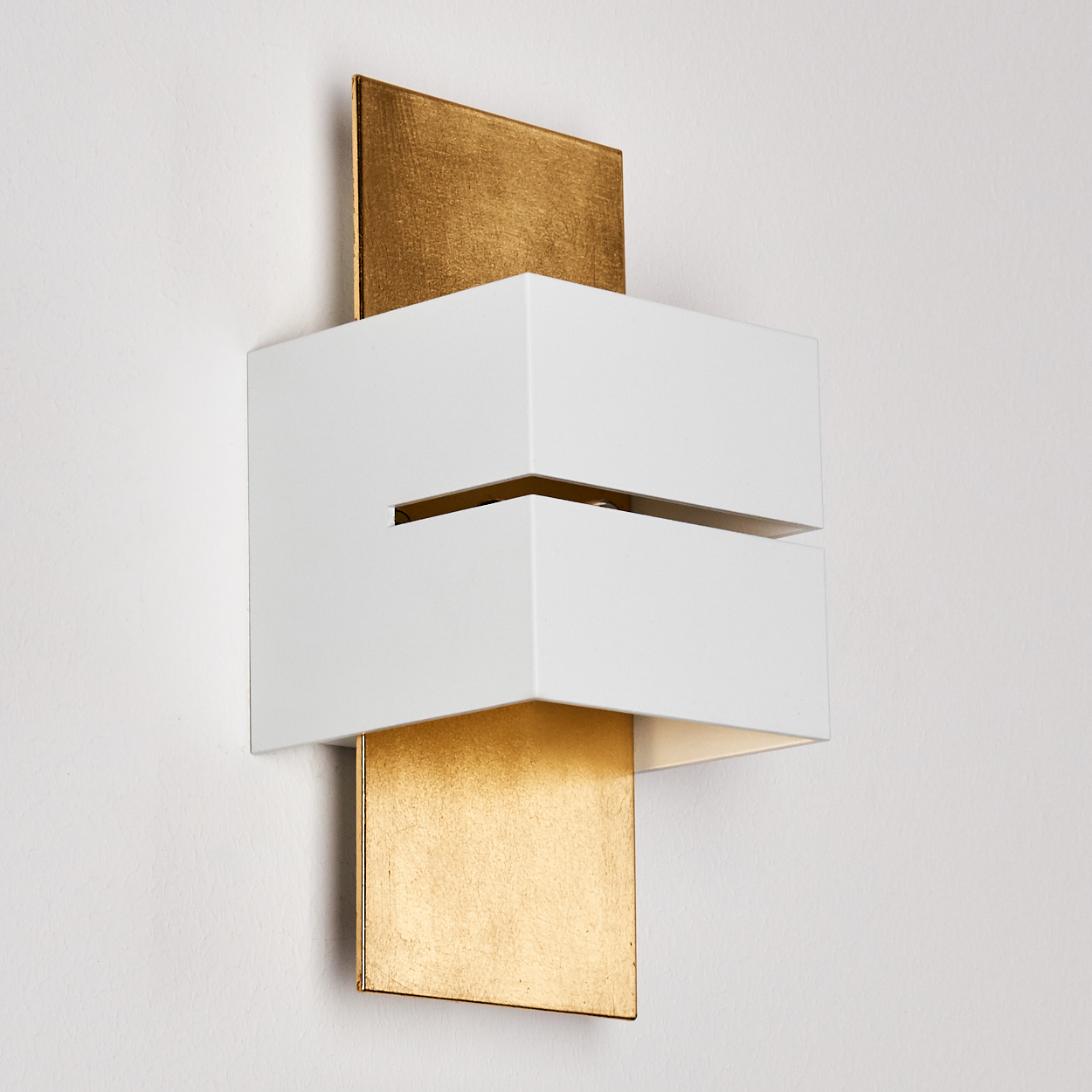 Eine moderne Wandlampe. Sie hat einen weißen, rechteckigen Schirm über einem goldenen Rechteck. Die Lampe ist an einer weißen Wand befestigt.