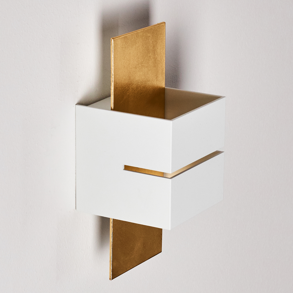 Ein modernes, geometrisches Wanddekor. Es hat weiße, kastenförmige Elemente mit goldenen Akzenten und goldenen Paneelen. Vor einer weißen Wand.