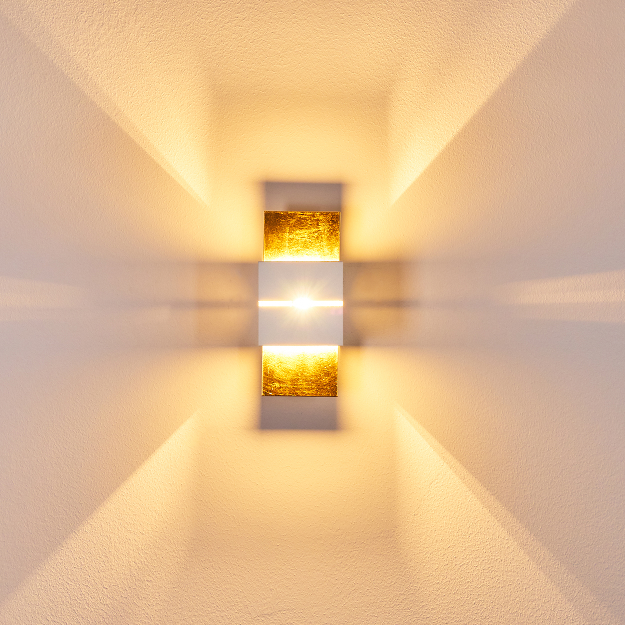 Eine moderne Wandleuchte mit Gold- und Weißelementen. Das Licht scheint auf die strukturierte Wand und wirft Schatten.