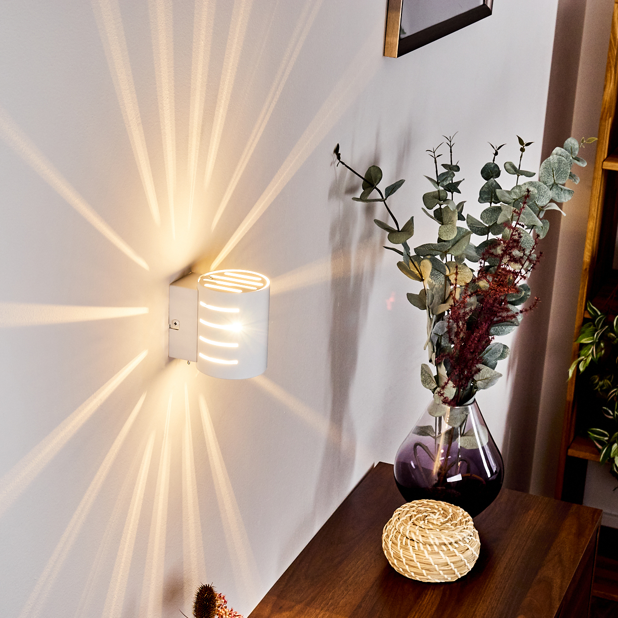Eine weiße Wandlampe strahlt Lichtstrahlen auf die Wand. Eine Vase mit Blumen und ein Korb stehen auf einem Holztisch.