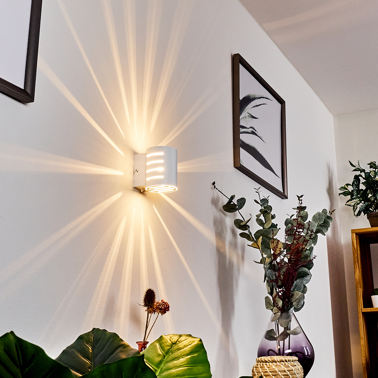 Weiße Wandlampe, die Lichtstrahlen aussendet. Kunstwerk an der Wand, Vase mit Blumen und Pflanzen im Vordergrund.